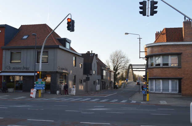 Stadtstraßenkreuzung mit Ampel, umgeben von Gebäuden, Fußgängern, Fahrrädern, Bäumen, Schildern, Laternenpfählen, einem Bogen im Hintergrund und einem klaren blauen Himmel.