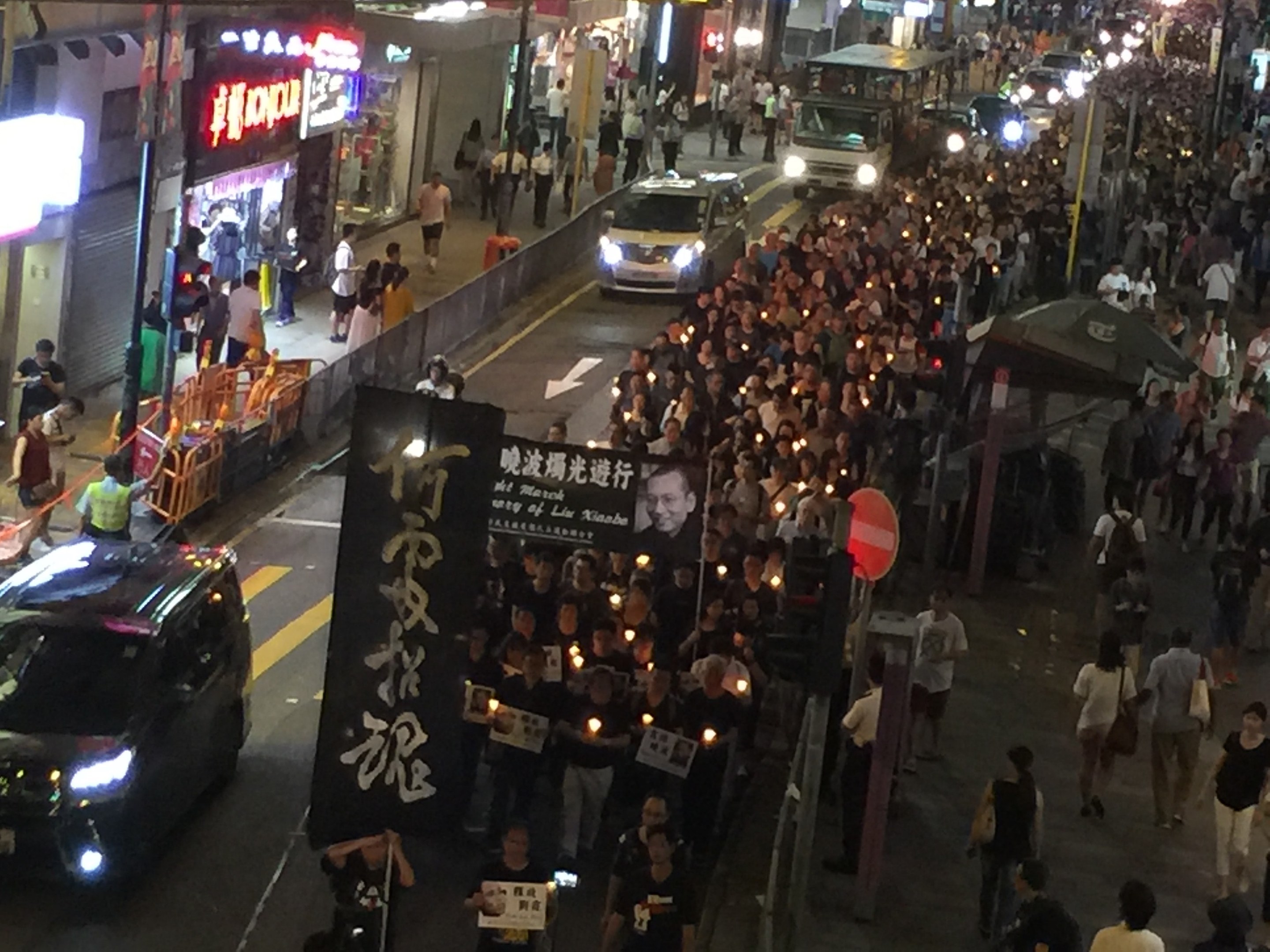 Eine große Gruppe von Menschen marschiert nachts auf einer Straße während einer Demonstration in Hong Kong, hält Schilder und Banner, mit Fahrzeugen, Straßenlaternen und Gebäuden im Hintergrund.