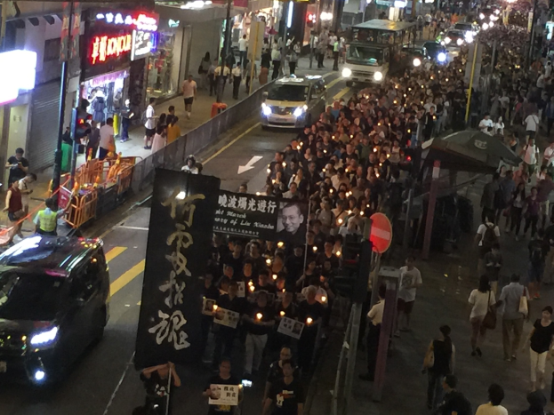 Eine große Gruppe von Menschen marschiert nachts auf einer Straße während einer Demonstration in Hong Kong, hält Schilder und Banner, mit Fahrzeugen, Straßenlaternen und Gebäuden im Hintergrund.
