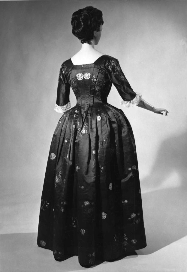 Eine Frau in einem 1950er-Vollkleiddress mit engem Oberteil und vollem Rock steht in einem Schwarz-Weiß-Foto, Hände verschränkt, trägt schwarze Pumps und eine klassische Hochsteckfrisur mit dezentem Make-up.