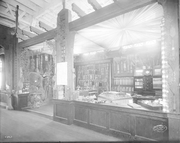 Ein altes Schwarz-Weiß-Foto einer Apotheke mit einer Theke, auf der verschiedene Gegenstände ausgebreitet sind und Regalen im Hintergrund, die mit Flaschen und anderen Gegenständen gefüllt sind.
