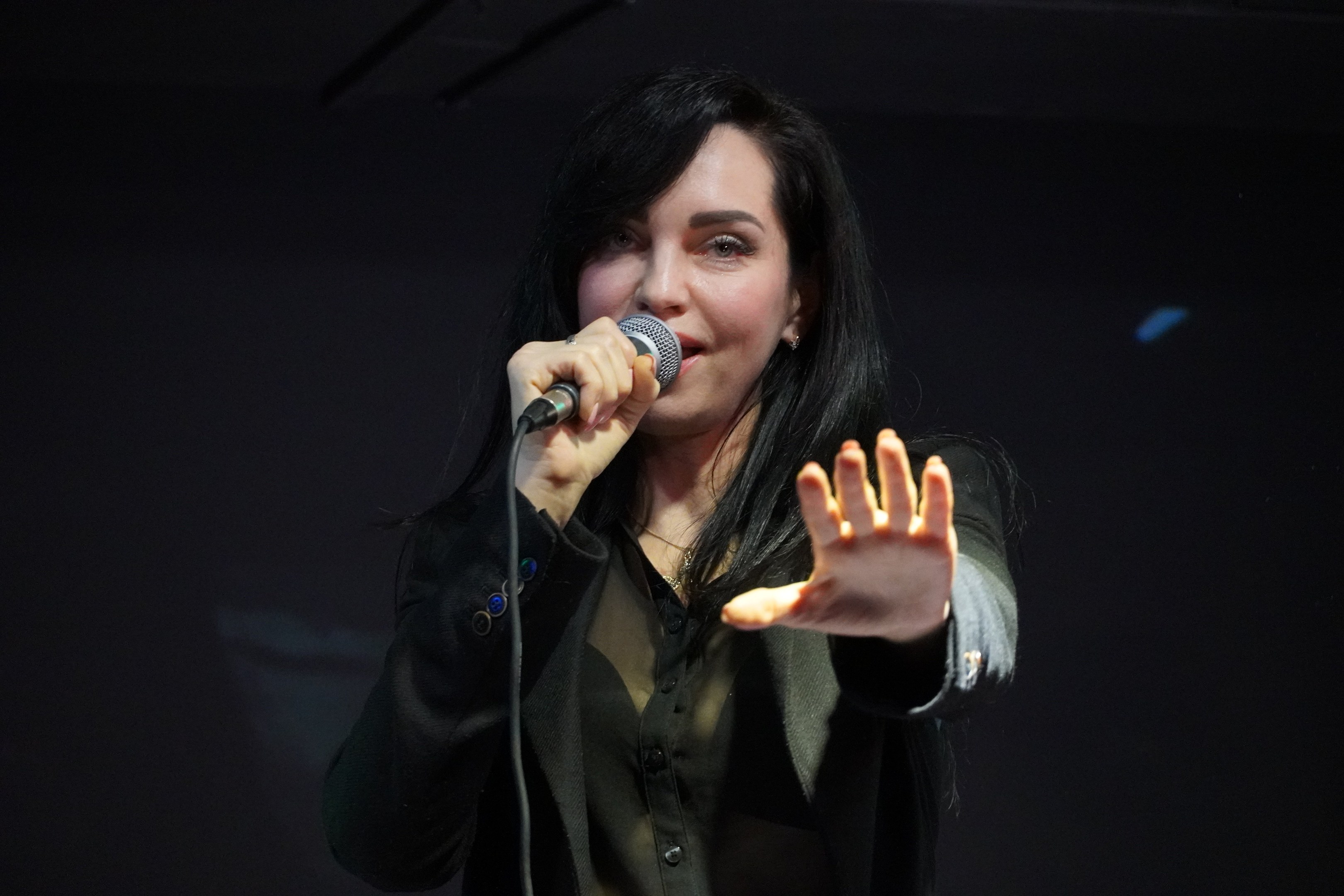 Frau singt leidenschaftlich in ein Mikrofon auf einer hell erleuchteten Bühne, trägt ein schwarzes Kleid und hat ihr Haar zu einem Knoten gebunden, die Augen geschlossen und einen Ausdruck voller Emotion.