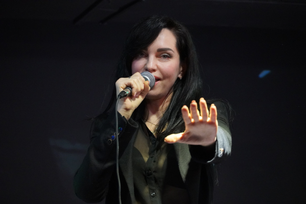 Frau singt leidenschaftlich in ein Mikrofon auf einer hell erleuchteten Bühne, trägt ein schwarzes Kleid und hat ihr Haar zu einem Knoten gebunden, die Augen geschlossen und einen Ausdruck voller Emotion.