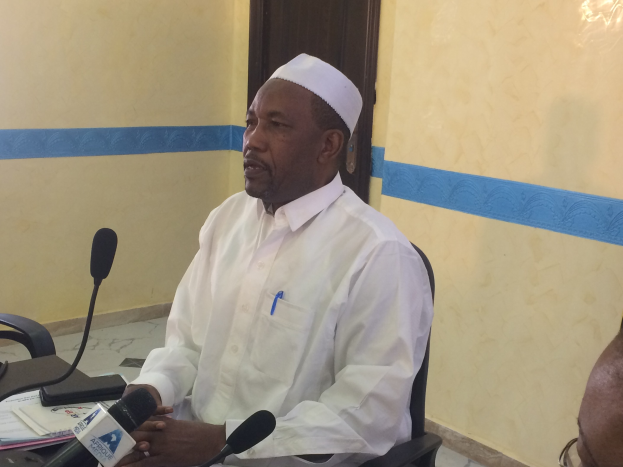 Ein Mann in einem weißen Gewand und einer Mütze sitzt an einem Tisch mit Mikrofonen, spricht, während Papiere und Gegenstände auf dem Tisch sichtbar sind, mit einer anderen Person mit Brille zu seiner Rechten, in einem Raum mit einer Wand und einer Tür dahinter.