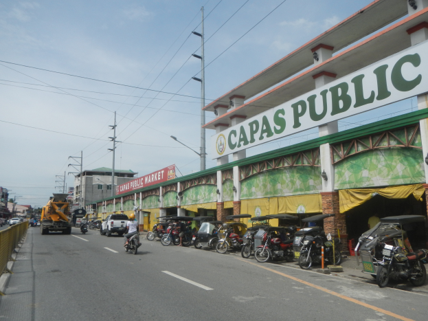 Eine belebte Stadtstraße mit Fahrzeugen, einem Geländer auf der linken Seite, Gebäuden, Pfählen, Lampen, Schildern und einigen Menschen unter einem bewölkten Himmel, mit einem Gebäude mit der Aufschrift "Capas Public".