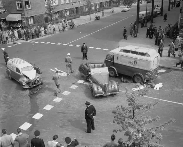 Eine Schwarz-Weiß-Szene eines Autounfalls an einer Straßenkreuzung mit mehreren Fahrzeugen, darunter einem Van, einer Gruppe von Menschen drumherum und im Hintergrund Gebäuden, Bäumen, Polen und Texttafeln.