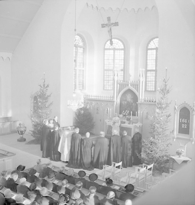 Eine Gruppe von Menschen in schwarz-weißer Kleidung, die auf Stühlen in einer Kirche sitzen und einem Chor zuhören, vor einem geschmückten Weihnachtsbaum, Kerzenständern, einem Kreuz an der Wand und Fenstern im Hintergrund.