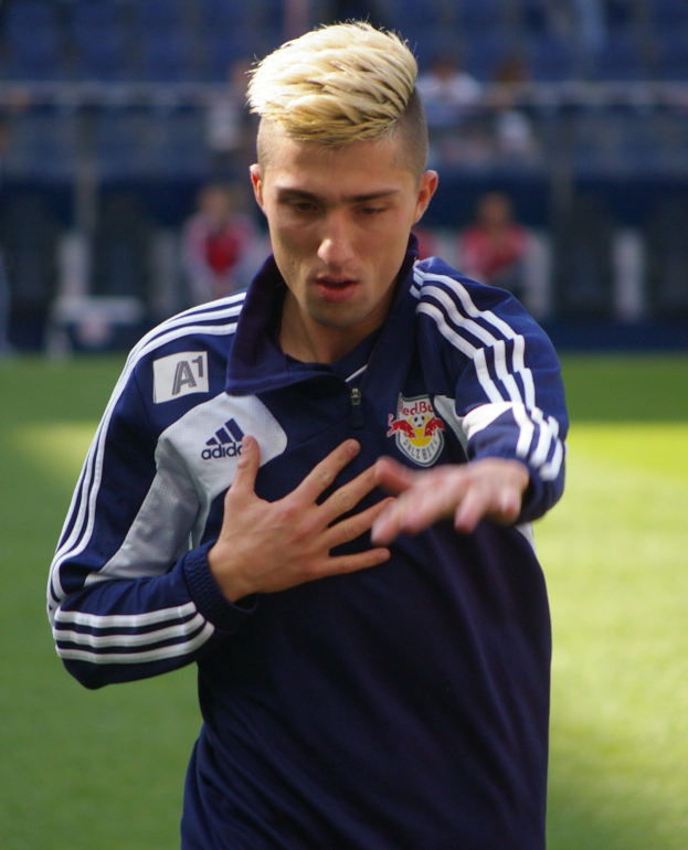 Ein junger Mann in einem blauen T-Shirt steht auf einem Fußballfeld und streckt die Arme aus, entschlossen als Flügelspieler.