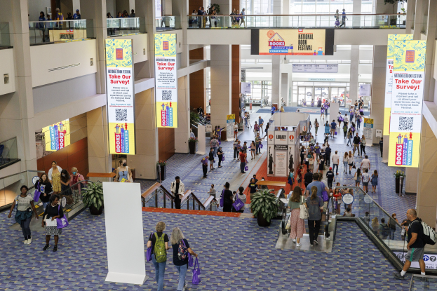 Eine große Gruppe von Menschen geht durch ein großes Gebäude mit Bannern, Topfpflanzen, Rolltreppen, Geländern, Säulen, bedruckten Tafeln, Glastüren und anderen Gegenständen, einige tragen Taschen, wahrscheinlich besuchen sie das National Book Festival.