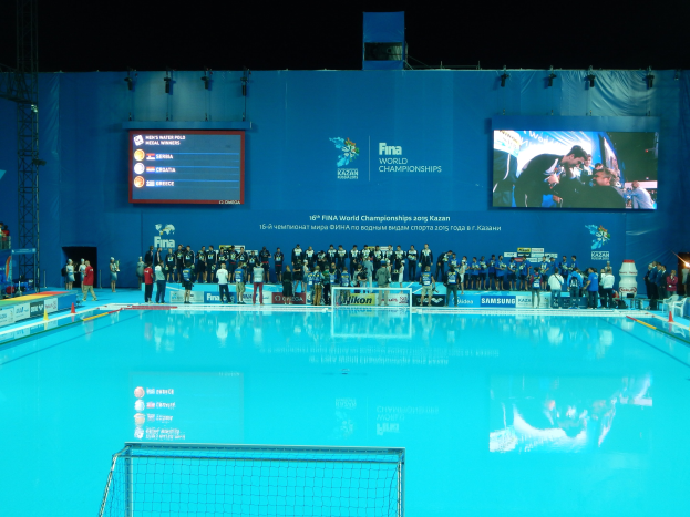 Eine Gruppe von Menschen um ein Hallenbad mit Netz am Boden herumstehend, mit einer Wand im Hintergrund, die "FINA World Championships 2015 Kazan"-Text und Logos anzeigt.