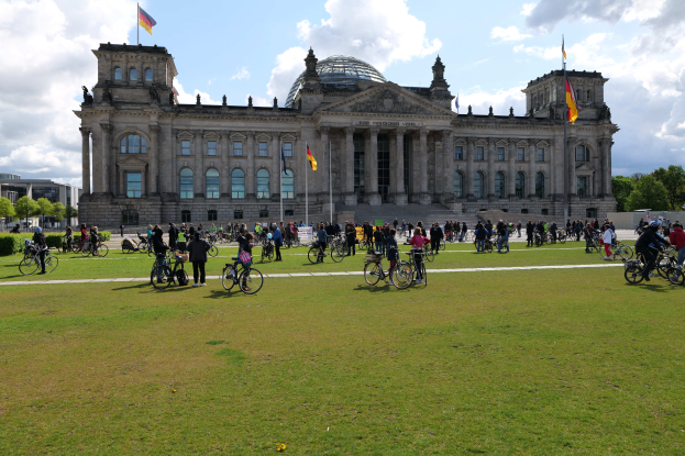 Eine Gruppe von Menschen, die Fahrr├Ąder vor dem Reichstaggeb├Ąude in Berlin, Deutschland fahren, das mit Fenstern, S├Ąulen und Flaggen mit Masten geschm├╝ckt ist, umgeben von B├Ąumen und Pflanzen, mit einem bew├Âlktem Himmel und grasbedecktem Boden.