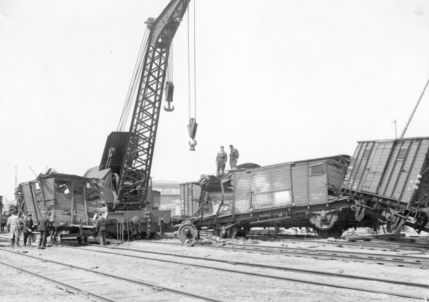 Schwarzes und weißes Bild einer Zugkatastrophe mit einem Kran, der ein Waggon auf die Schienen hebt, mehrere Personen in der Nähe und im Hintergrund Gebäude und einen klaren Himmel.