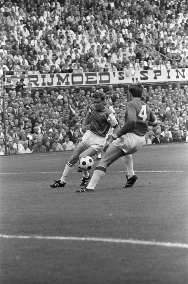 Schwarzes und weißes Foto von zwei Männern, die auf einem Feld Fußball spielen, mit Zuschauern im Hintergrund und einer Tafel mit der Aufschrift "Bobby Charlton im Einsatz für Manchester United."