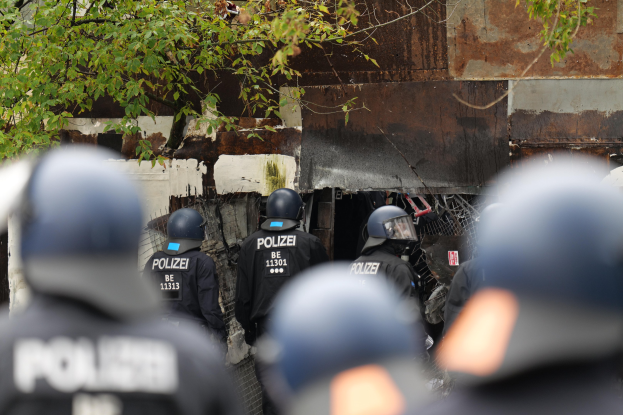 Gruppe von Polizisten in schwarzen Uniformen und Helmen vor einem durch Maschendrahtzaun umgebenen Geb├Ąude mit B├Ąumen im Hintergrund.
