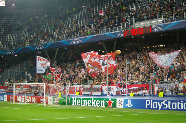 Ein Fussballspiel wird in einem hell erleuchteten Stadion mit einer grossen Zuschauermenge, grünem Rasen, einem Torpfosten mit Bannern und einem Bildschirm in der linken oberen Ecke gespielt.