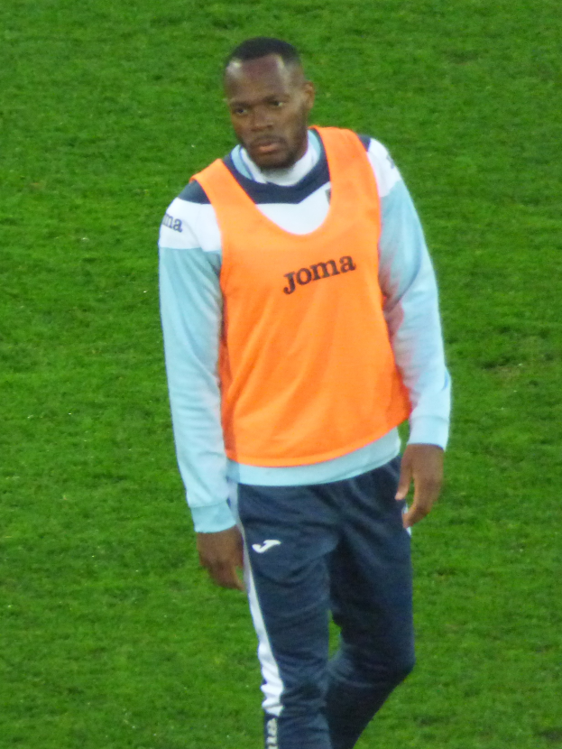 Ein Mann in einem orangen Hemd und blauen Hosen geht über ein grünes Feld und sieht entschlossen aus, wobei er wie ein Fußballspieler aussieht.