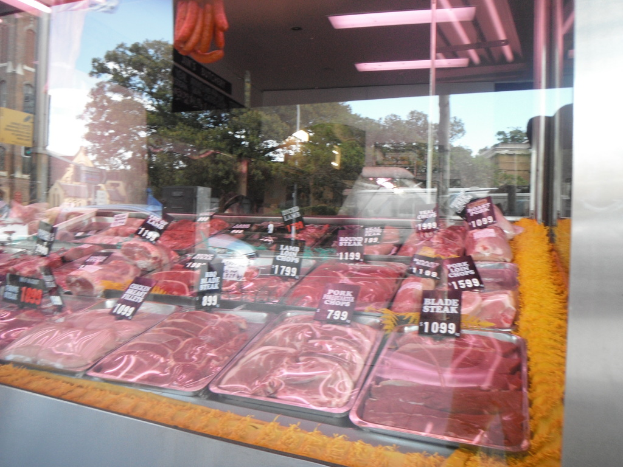 Eine Glasvitrine in einem Fleischgeschäft mit Tabletts voller Fleisch und Preisschildern, mit Gebäuden und Bäumen im Hintergrund und der Decke des Raums oben im Bild.