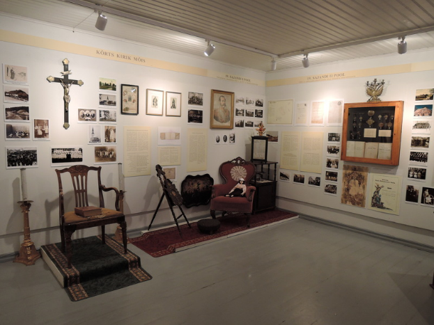 Ein Museumsraum mit Stühlen, Teppichen, Kerzenständern, einem Kreuzsymbol an der Wand, Fotorahmen, anderen Gegenständen auf Tischen und Deckenleuchten.