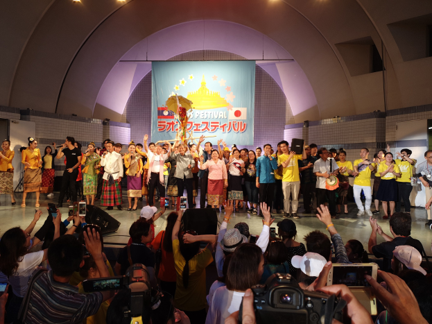 Eine Gruppe von Menschen auf einer Bühne mit Kameras, Handys und Musikinstrumenten während einer scheinbar festivalartigen Eröffnungszeremonie, mit einem Banner, Wänden und Deckenlampen im Hintergrund.