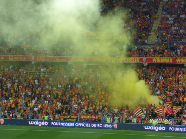 Ein Fußballstadion voller Menschen, einige halten Fahnen und Banner, andere stehen oder sitzen, mit einer Wolke aus Rauch in der Luft.