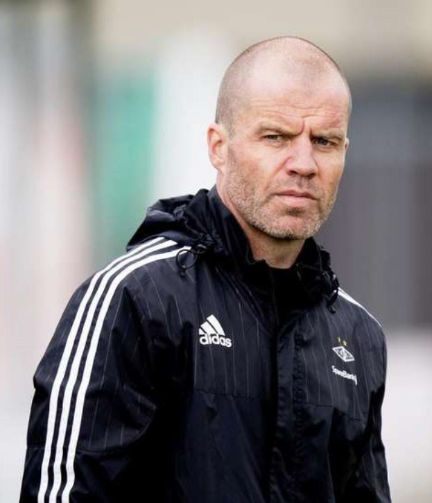 Ein kahlköpfiger Mann in einer blauen Jacke mit weißen Streifen und weißem Kragen steht auf einem Fußballfeld, die Hände verschränkt und einen ernsten Gesichtsausdruck.