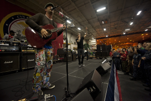 Ein als Chris Stapleton identifizierter Mann spielt eine E-Gitarre auf einer Bühne, trägt eine Mütze, hat ein Mikrofon, Lautsprecher, eine Fahne und andere Instrumente in der Nähe und tritt vor einem Publikum unter Deckenlampen mit einem Banner an der Wand hinter ihm auf.