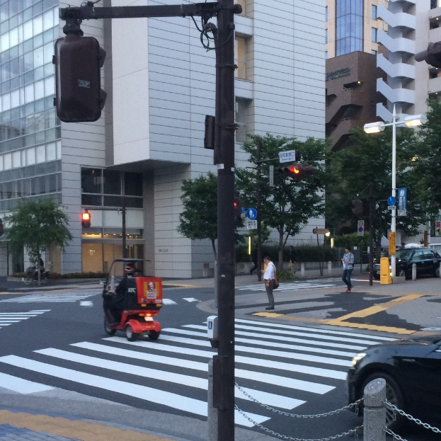 Ein E-Scooter auf einer Straße mit Personen im Hintergrund, eine Ampel im Vordergrund und Gebäude mit Bäumen dahinter.