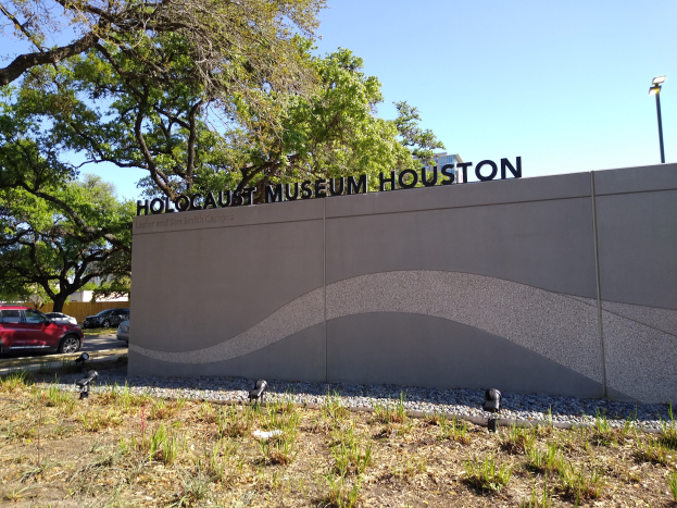 Außenansicht des Houston Holocaust Museums mit einer Textwand, Gedenksteine, Grünanlage, Straße mit Fahrzeugen, einer Straßenlaterne und einem bewölkten Himmel.