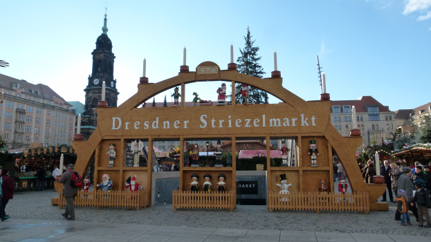 Ein lebendiger Weihnachtsmarkt in Dresden, Deutschland, mit Menschen, die herumlaufen, einige mit Taschen, einem Bogen mit Text, Tänzen, Gebäuden mit Fenstern und einem bewölktem Himmel.