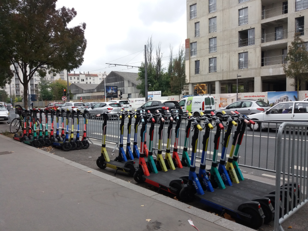 Eine Reihe von Elektro-Scootern, die auf der Straßenseite geparkt sind, mit einem Geländer auf der rechten Seite, Gebäude, Bäume, Masten, Verkehrszeichen und Schilder im Hintergrund unter einem klaren blauen Himmel.