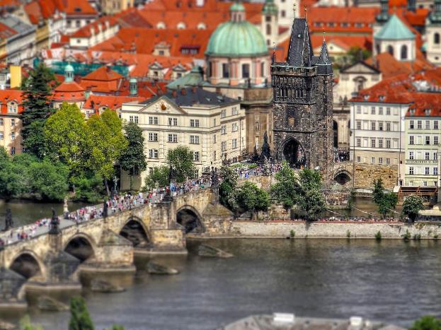 Eine Stadtansicht mit zahlreichen Gebäuden, einem prominenten Turm, Bäumen, Menschen, einem Fluss und einer Brücke, die den Fluss überquert.