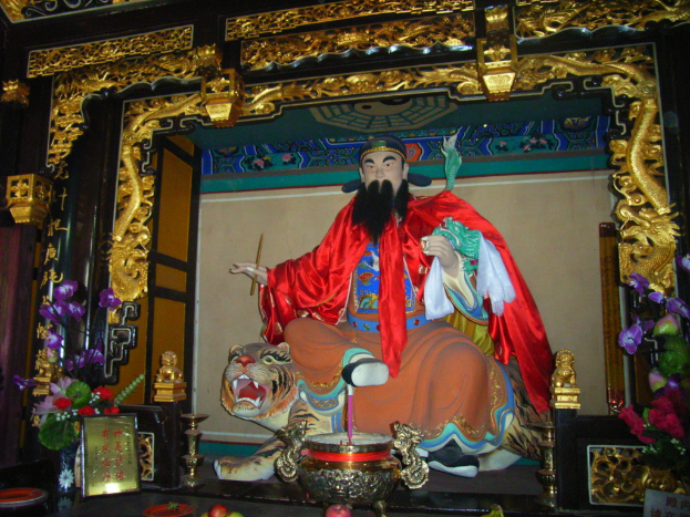 Eine Statue eines chinesischen Mönchs mit buntem Stoff auf einem Tiger sitzt unter einem Bogen mit goldenen Verzierungen, vorne ein Blumenkübel und in der Mitte eine Schüssel.