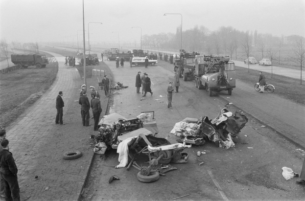 Eine Schwarz-Weiß-Szene eines Autounfalls am Straßenrand mit mehreren Fahrzeugen, darunter ein Landfahrzeug, und einer Gruppe von Menschen drumherum, mit Laternenmasten, Bäumen und Himmel im Hintergrund.
