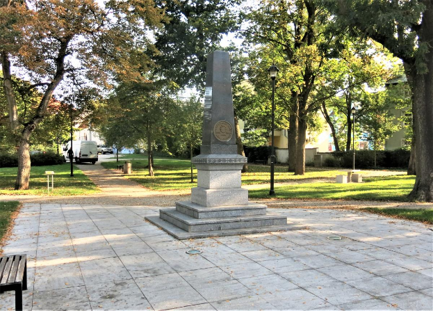 Ein Denkmal steht in einem Park mit einer Bank auf der linken Seite, umgeben von Bäumen, Laternenmasten und Fahrzeugen auf einer nahen Straße, mit Gebäuden und einem klaren blauen Himmel im Hintergrund.