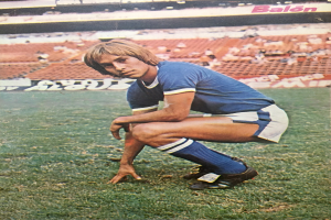 Ein Mann kniet auf einem Fußballfeld, umgeben von sitzenden Personen mit Schildern, die Text tragen und Sportschuhe tragen.