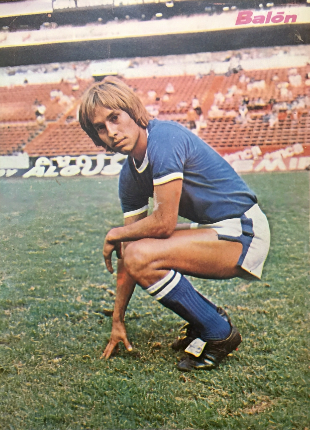 Ein Mann kniet auf einem Fußballfeld, umgeben von sitzenden Personen mit Schildern, die Text tragen und Sportschuhe tragen.