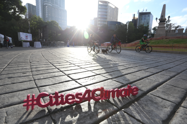 Eine Gruppe von Menschen, die auf Fahrrädern eine Straße entlangfahren, mit der Aufschrift "Cities4Climate" darauf, mit Gebäuden, Bäumen, Pfählen, Laternen, Bannern, Geländern, Gras, einer Statue und Wolken im Hintergrund.