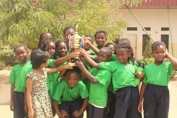 Eine Gruppe von Kindern hält in einer Schulumgebung mit Bäumen und einem Gebäude im Hintergrund einen Pokal