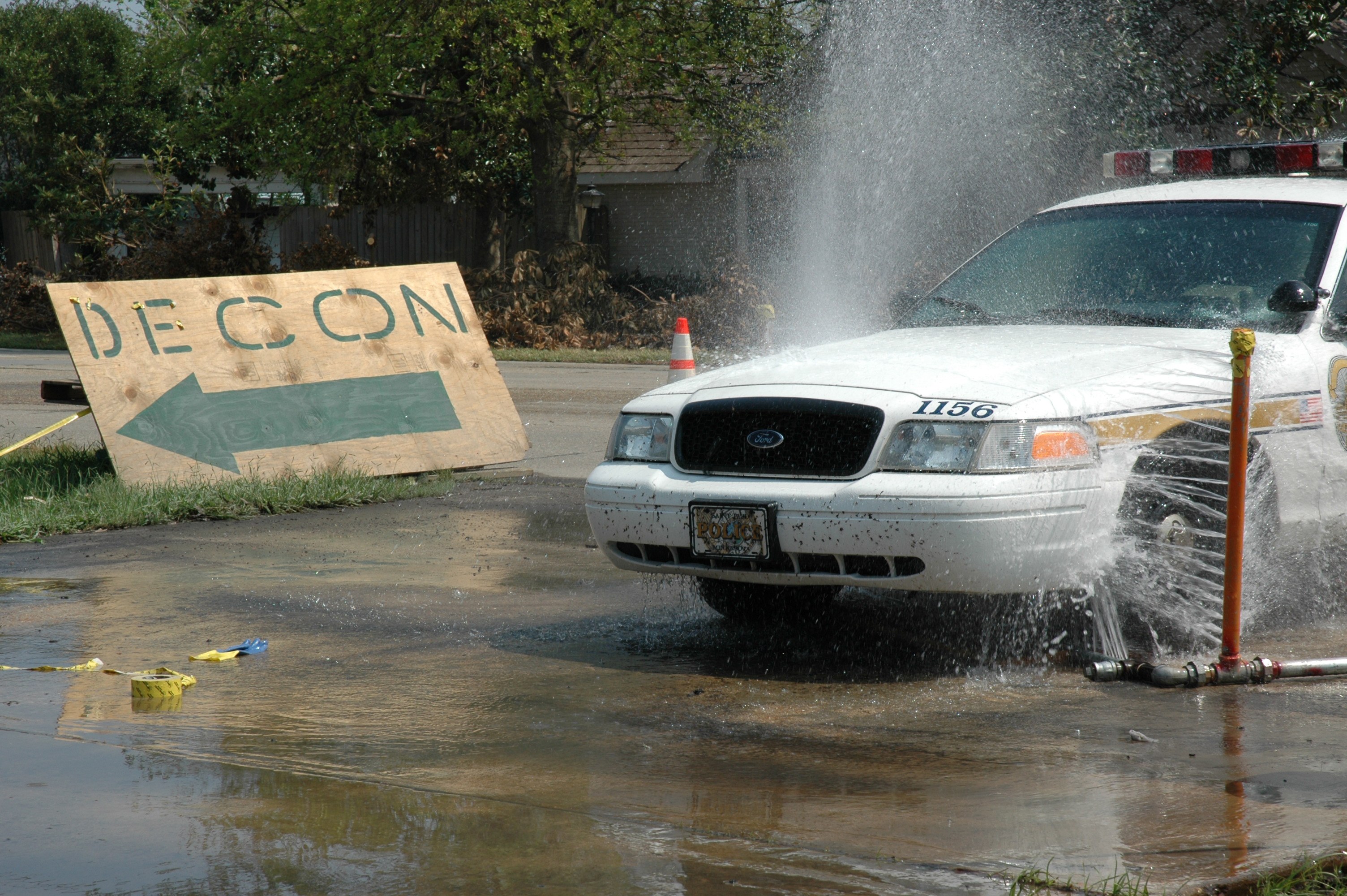 Ein Polizeiwagen fährt durch eine Pfütze, mit einer Tafel, einem Verkehrskegel und anderen Gegenständen am Boden; Bäume, Gebäude und ein klarer blauer Himmel sind im Hintergrund zu sehen.