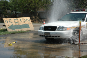Ein Polizeiwagen fährt durch eine Pfütze, mit einer Tafel, einem Verkehrskegel und anderen Gegenständen am Boden; Bäume, Gebäude und ein klarer blauer Himmel sind im Hintergrund zu sehen.