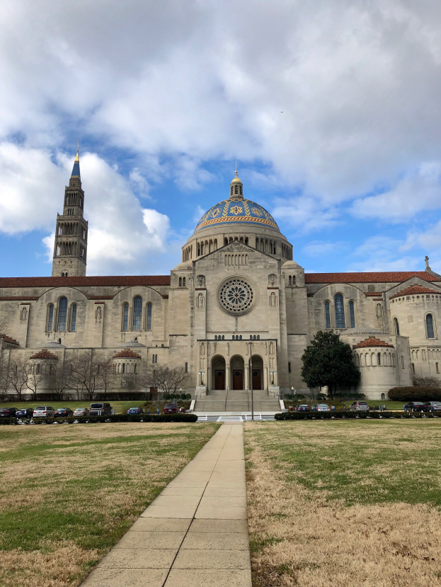 Die Basilika des Heiligen Herzens Jesu in St. Louis, Missouri, ein großes Gebäude mit Fenstern, Türen und Säulen, umgeben von einem Weg, Gras, Bäumen, Fahrzeugen, Pfählen und einem bewölkten Himmel.