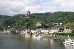 Ein malerischer Blick auf den Rhein in Deutschland mit einer Burg auf einem Hügel, Booten auf dem Fluss und Fahrzeugen auf einer näheren Straße sowie einem bewölktem Himmel.