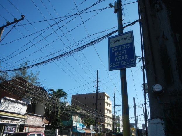 Eine Stadtstraße mit Autos, Strommasten mit Drähten, Gebäuden, Bäumen und Namensschildern, mit einem Schild an einem Mast, auf dem steht "Fahrer müssen Sitzgurte tragen" und einem sichtbaren Himmel im Hintergrund.