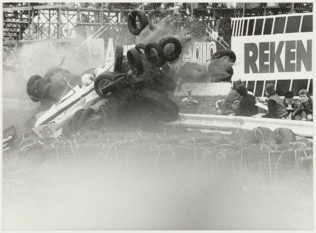 Schwarz-weiß-Foto eines verunglückten Rennwagens umgeben von Reifen, mit einer Gruppe von Menschen auf der rechten Seite und einer Tafel mit Text im Hintergrund.