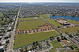 Luftbild einer Stadt mit Gebäuden, Bäumen, Gras, Straßen mit Fahrzeugen, Wasser und Wolken, das einen großen zentralen Platz zeigt, der als Parkgebiet vorgeschlagen ist.
