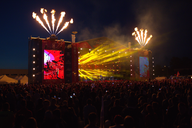 Eine große Menschenmenge auf einem Musikfestival mit einer hell erleuchteten Bühne mit Musikern und Transparenten, Zelte links daneben und Feuerwerk am dunklen Himmel.