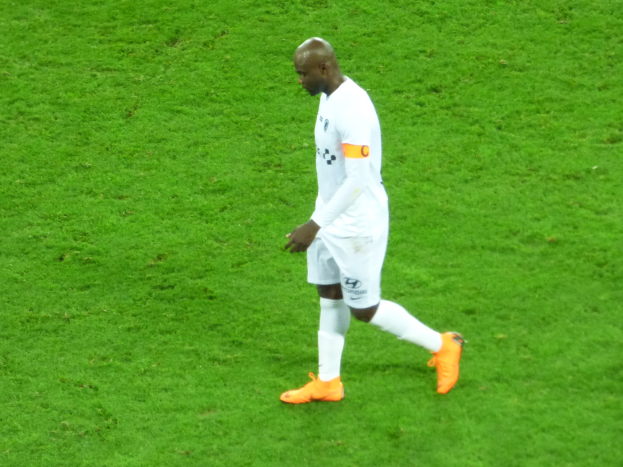 Ein Fussballspieler in einem weissen T-Shirt, weissen Shorts und orangenen Schuhen läuft über ein saftiges grünes Feld.