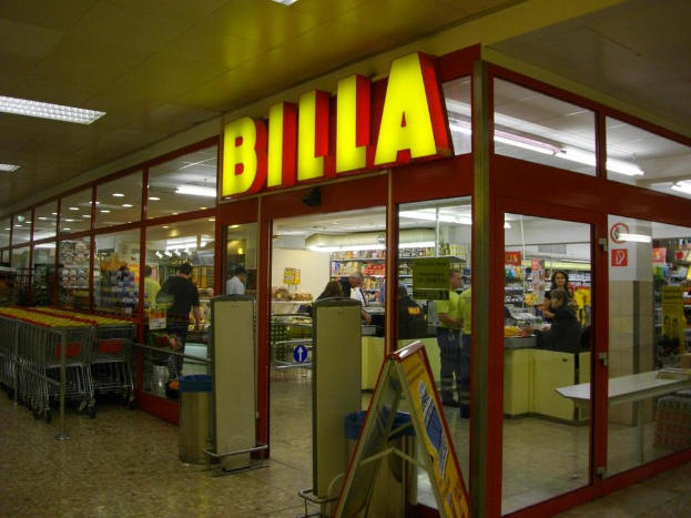 Ein Markt mit einem Eingang in der Mitte, Mülltonnen daneben, einer Reihe von Einkaufswagen links, Deckenbeleuchtung und einigen Menschen, die Produkte betrachten.