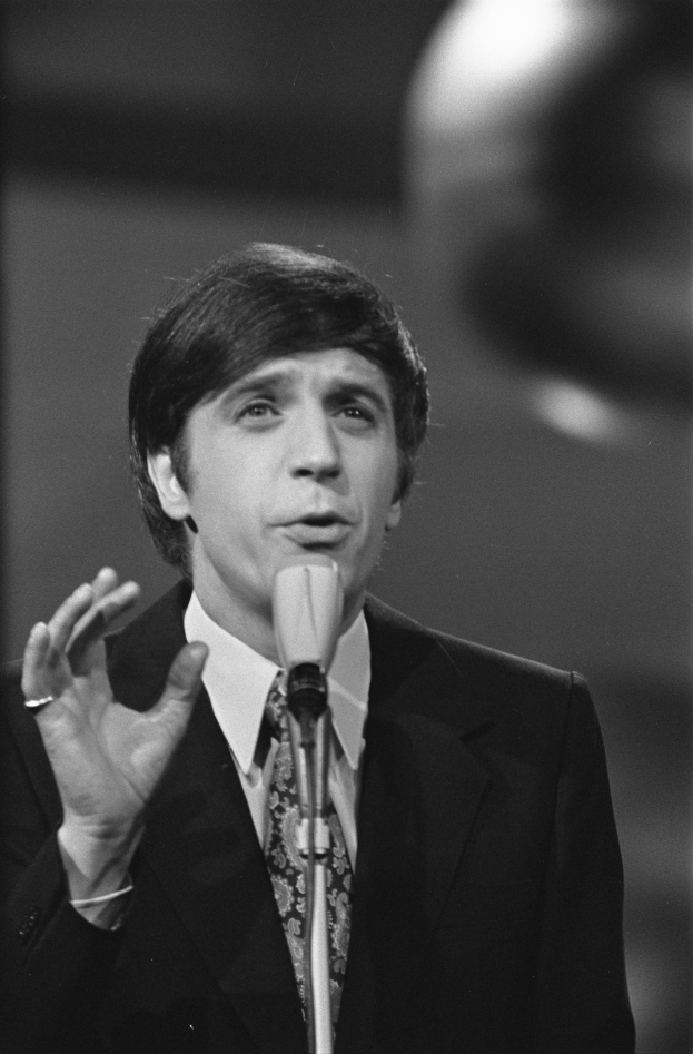 Ein Mann in einem schwarzen Anzug, weißem Hemd und gemusterter Krawatte spricht in ein Mikrofon und hat einen ernsten Gesichtsausdruck, sein Haar ist zurückgekämmt.