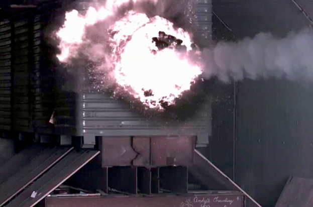 Eine große Explosion in der Mitte eines Gebäudes mit Rauch, einer Wand im Hintergrund und metallischen Gegenständen mit einer Tafel unten.